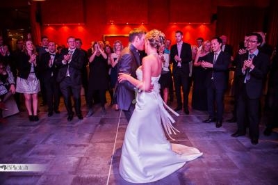 Un couple danse au centre d'une salle entouré d'invités applaudissant dans une ambiance festive et éclairage chaleureux.
