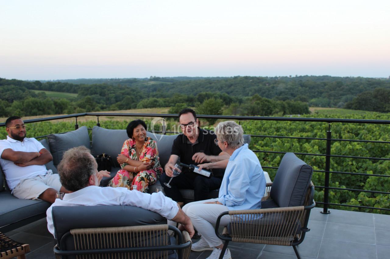 Un groupe de personnes discute assis sur une terrasse avec vue sur un paysage verdoyant au coucher du soleil.