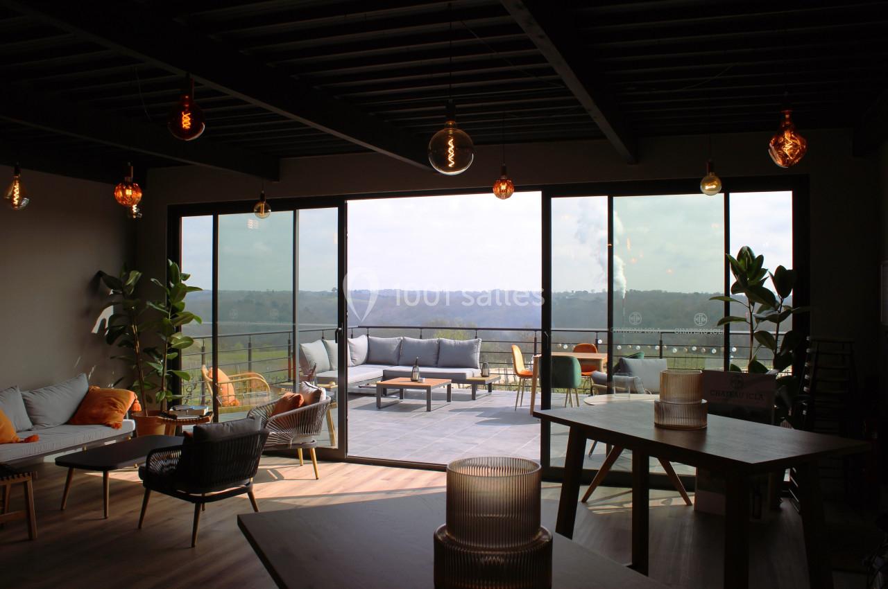 Salle lumineuse avec grandes baies vitrées donnant sur une terrasse aménagée et vue sur un paysage vallonné.
