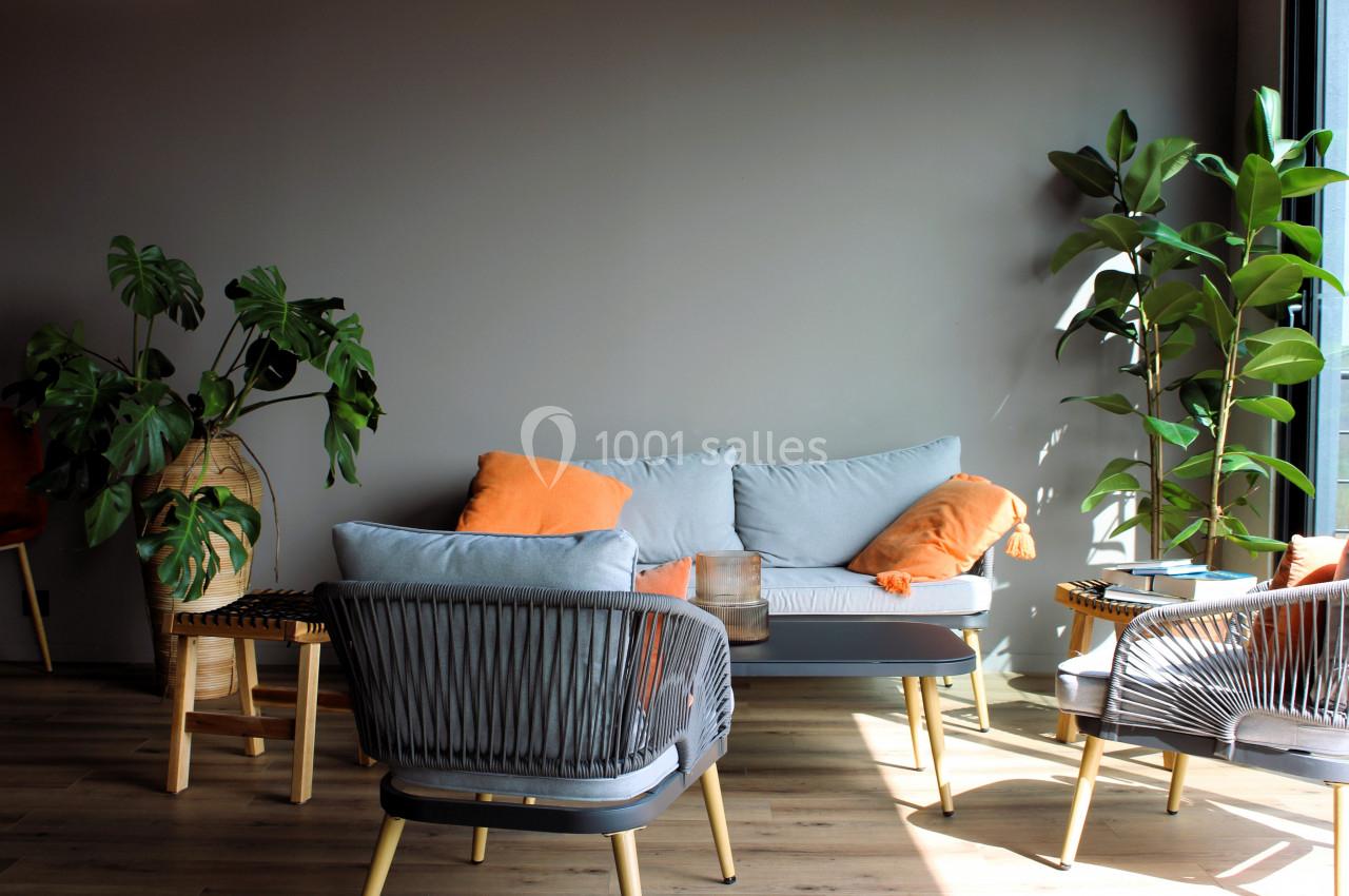 Salon lumineux avec canapé gris, coussins orange, plantes vertes en pots et fauteuils en rotin sur un sol en bois.