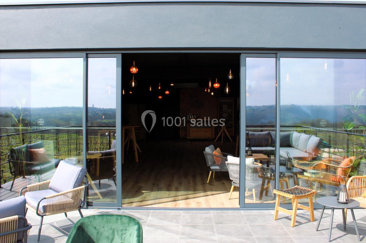 Vue d'une terrasse aménagée avec des fauteuils et une table, donnant sur une salle intérieure lumineuse et un paysage…