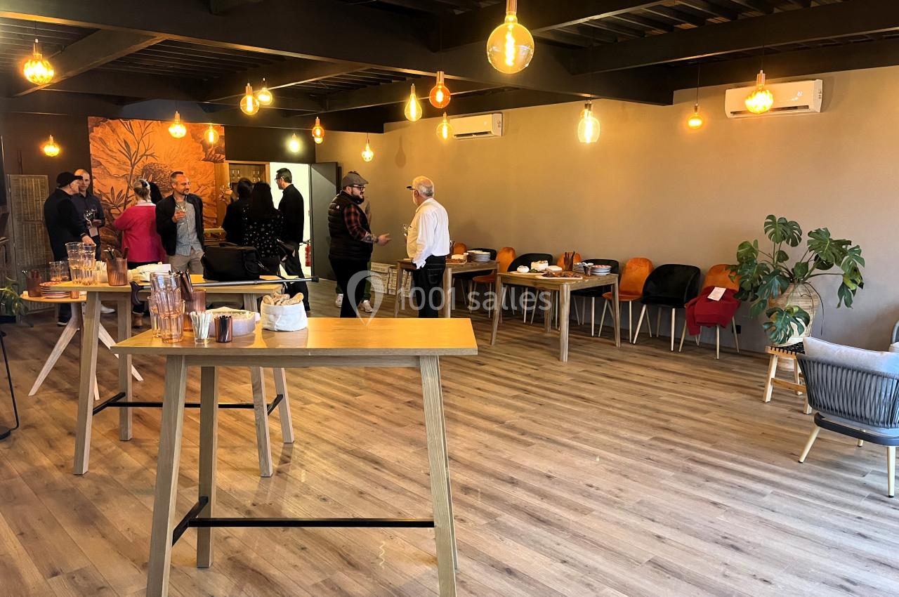 Salle lumineuse avec parquet, tables hautes, chaises colorées et personnes discutant lors d'un événement informel.