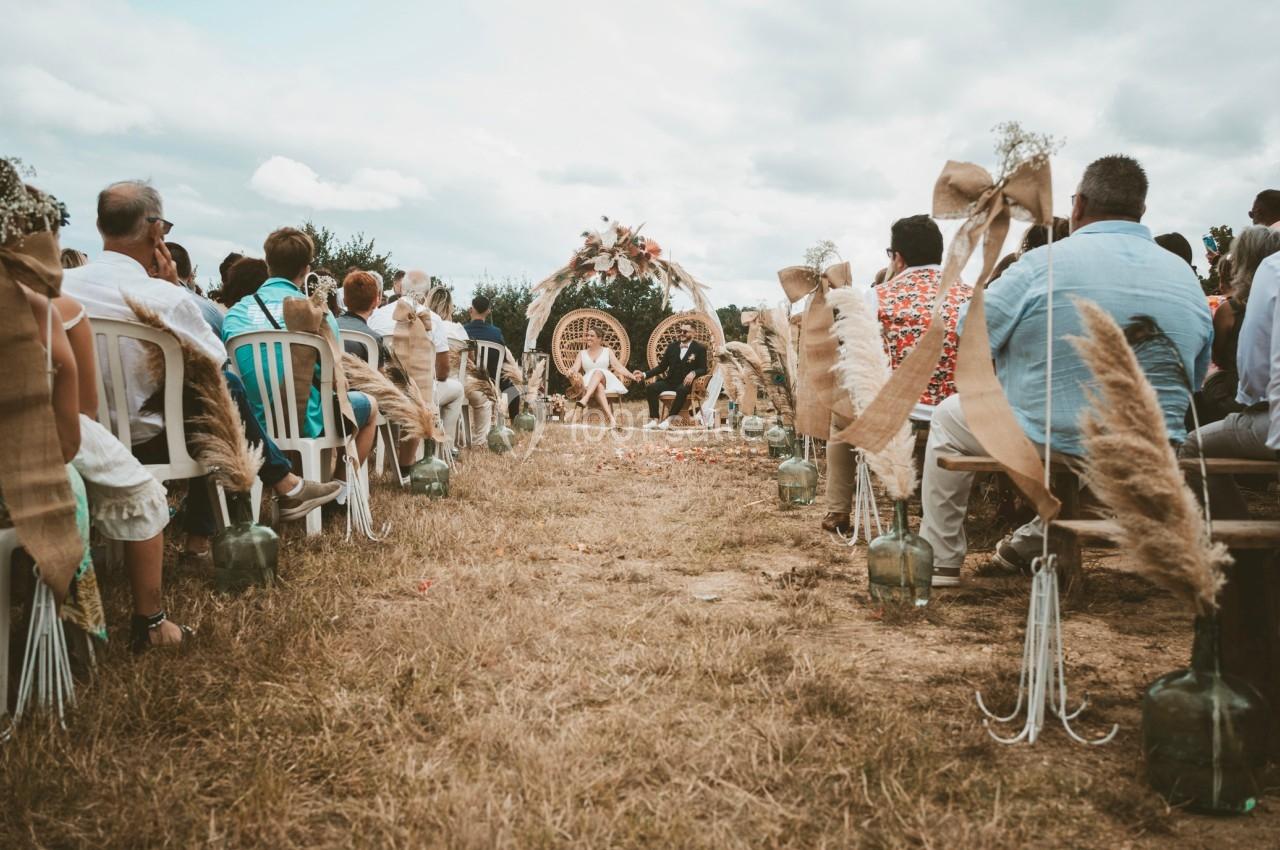 Cérémonie de mariage en plein air avec des invités assis, décor champêtre et arche ornée de fleurs au centre.