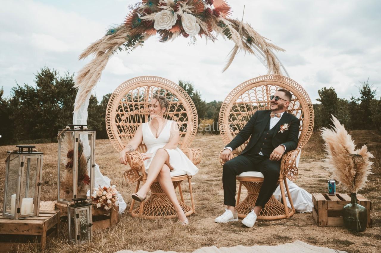 Un couple assis sur des fauteuils en rotin sous une arche décorée de fleurs et de pampas dans un décor champêtre.