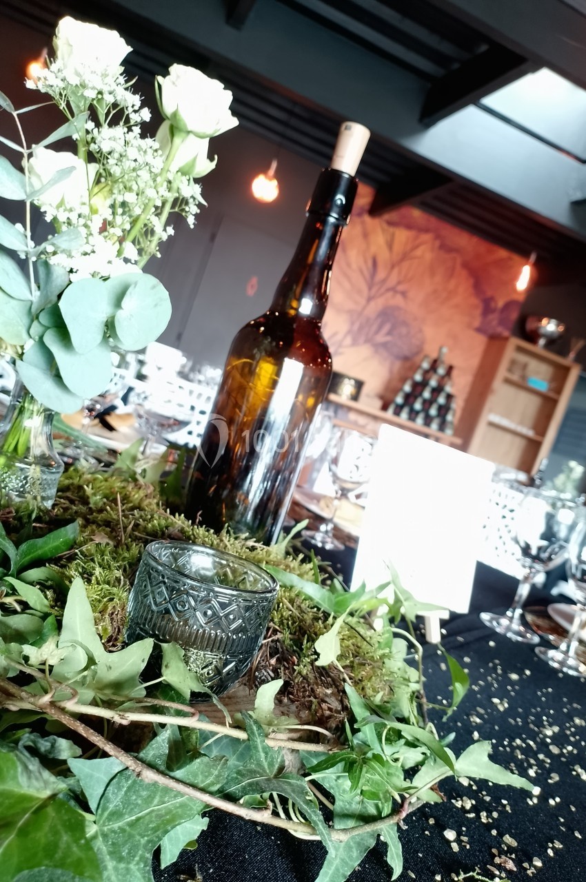 Location salle Saint-Genès-de-Lombaud (Gironde) - Domaine du Château Icla #25 Centre de table avec une bouteille en verre, feuillage, mousse et bougie, dans une salle décorée pour un événement.