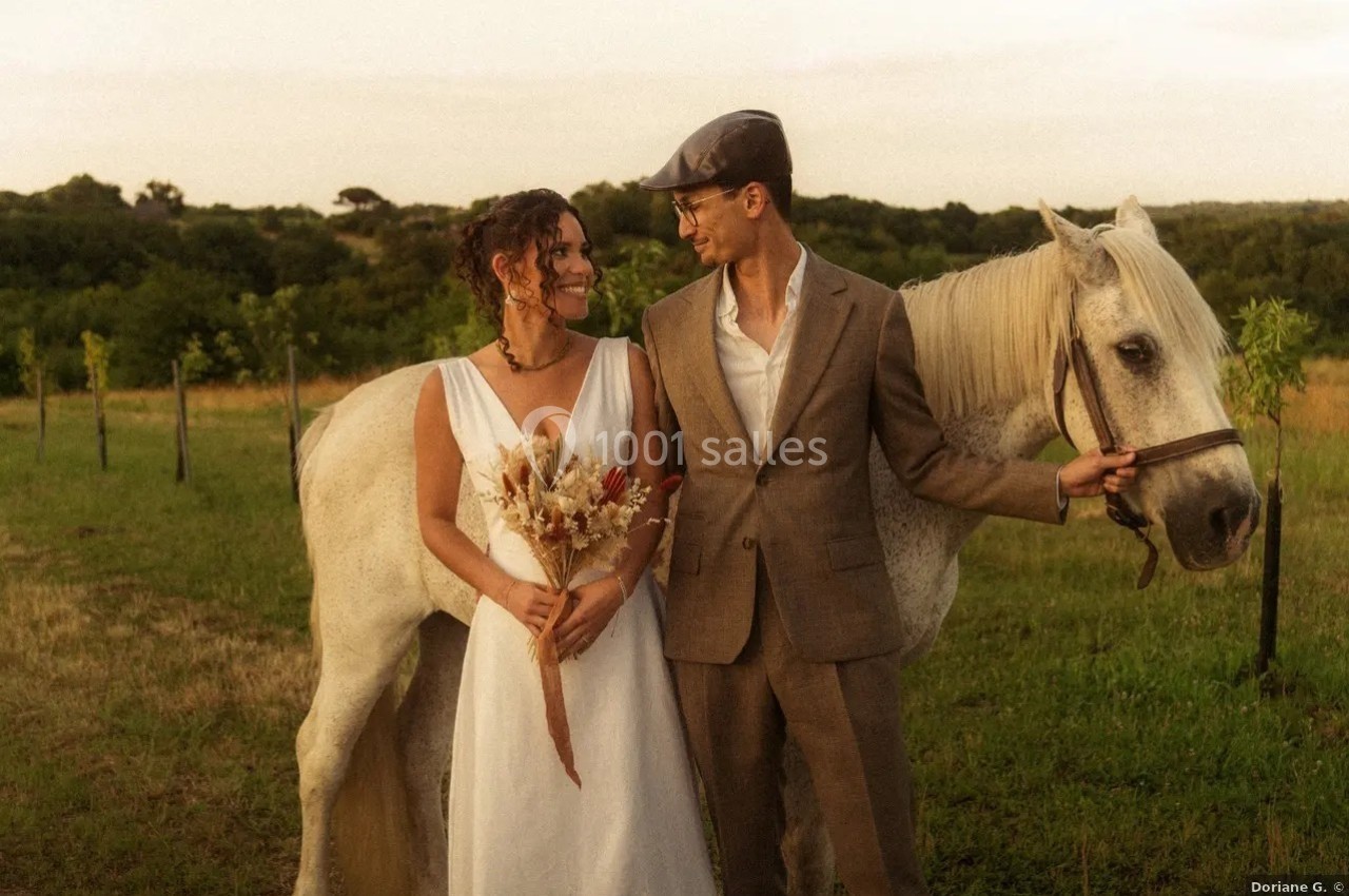 Location salle Saint-Genès-de-Lombaud (Gironde) - Domaine du Château Icla #38 Un couple souriant en tenue élégante se tient près d'un cheval blanc dans un champ verdoyant au coucher du soleil.