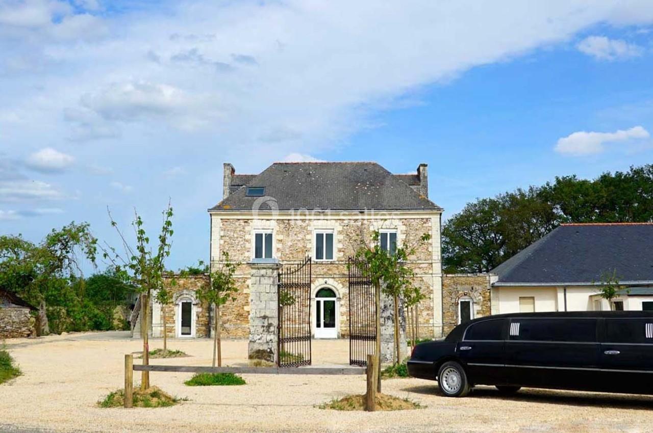Façade d'une maison en pierre avec portail en fer forgé, entourée d'arbres, et une limousine noire stationnée devant.