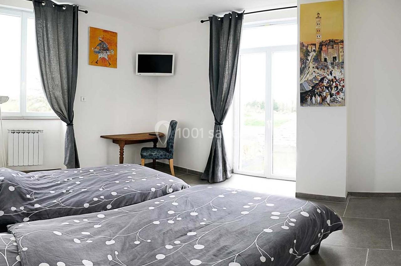 Chambre lumineuse avec deux lits simples, rideaux gris, bureau en bois, télévision murale et décorations murales colorées.