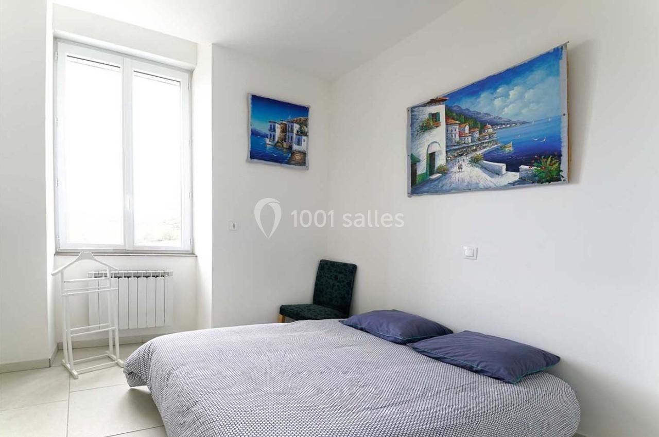 Chambre lumineuse avec un lit double, une chaise, un petit bureau blanc et des tableaux représentant des paysages côtiers.