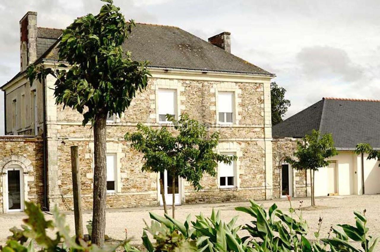 Maison en pierre avec toit en ardoise, entourée d'arbres et de végétation, sous un ciel partiellement nuageux.