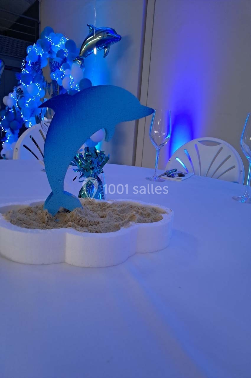 Centre de table avec un dauphin bleu en carton, posé sur du sable, entouré de décorations bleues et blanches.