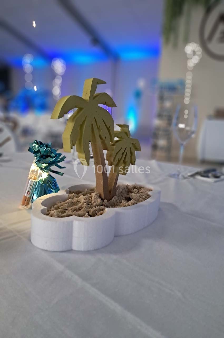 Centre de table avec des palmiers miniatures en bois plantés dans du sable, sur une nappe blanche.
