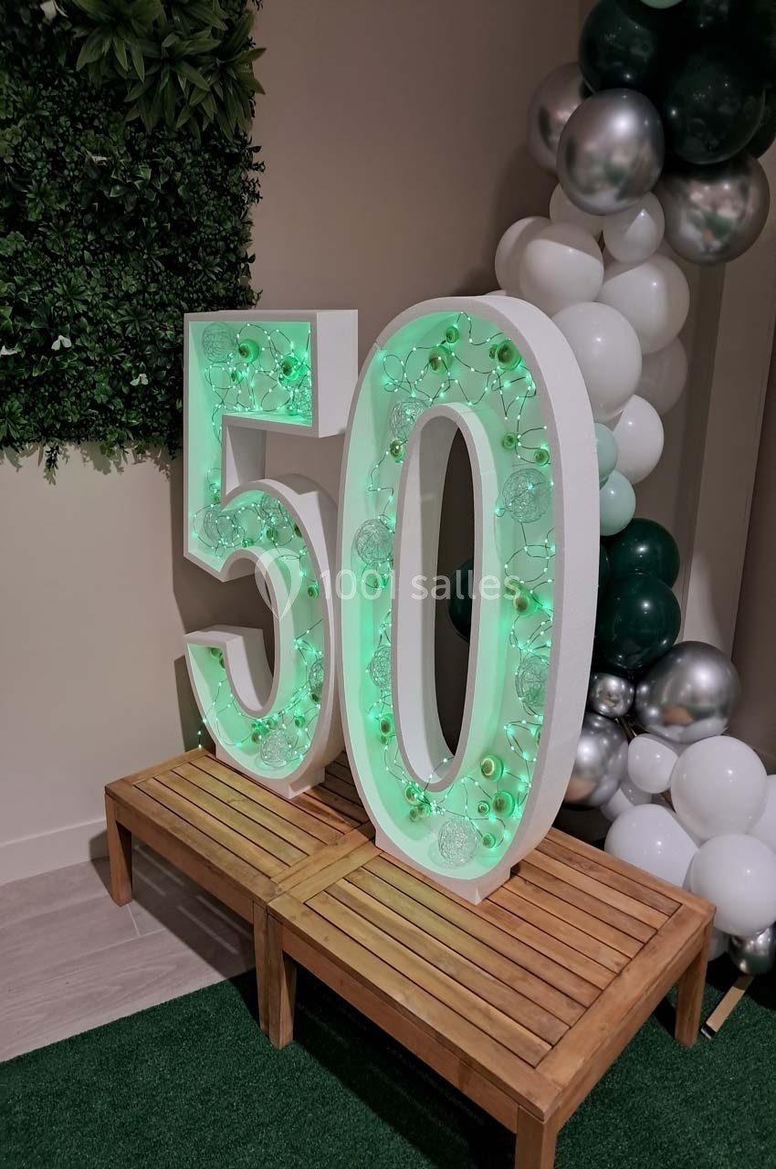 Chiffres ’50’ lumineux décorés de guirlandes vertes, posés sur un banc en bois, avec des ballons et un mur végétal en…
