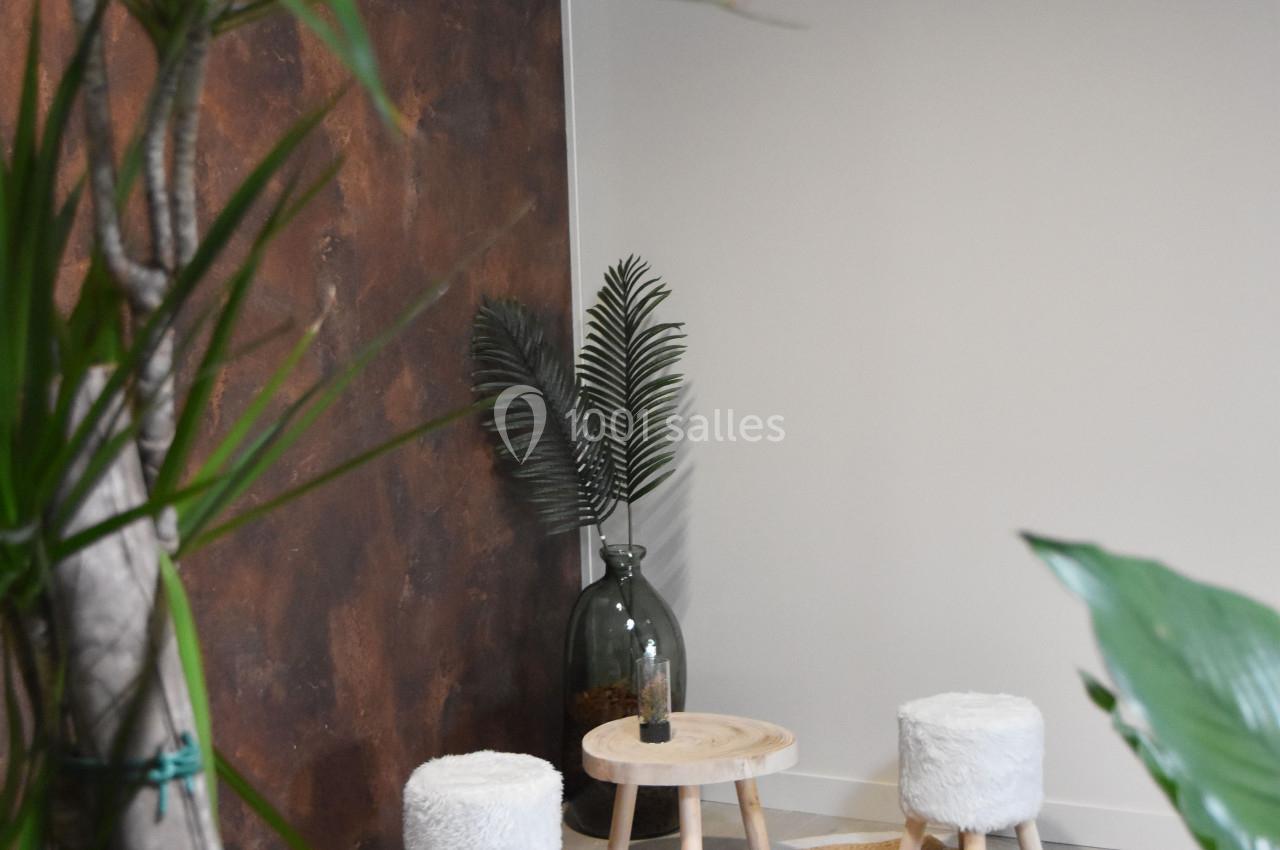 Deux tabourets blancs et une petite table en bois sur des tapis ronds, entourés de plantes dans un espace lumineux.