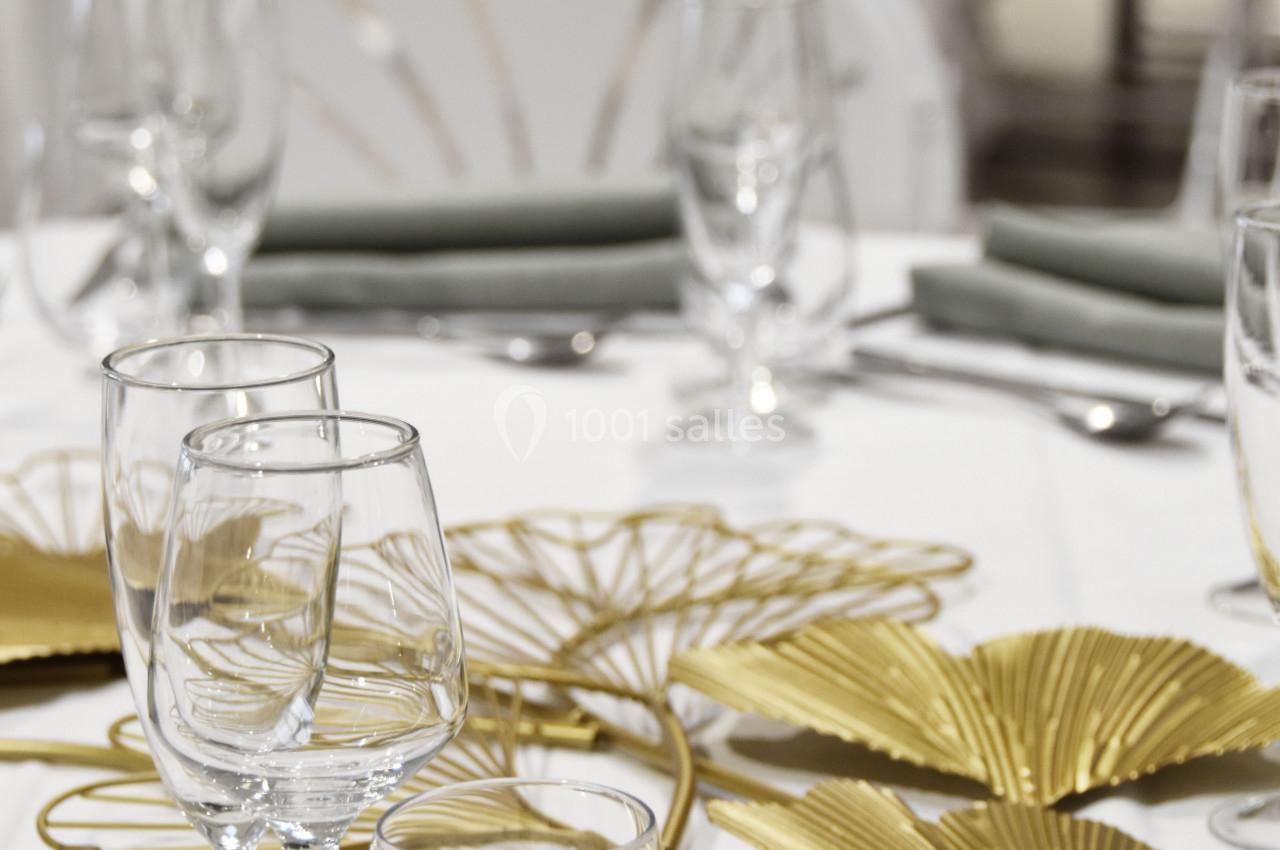 Table dressée avec nappes blanches, verres, couverts et décoration dorée en forme de feuilles.