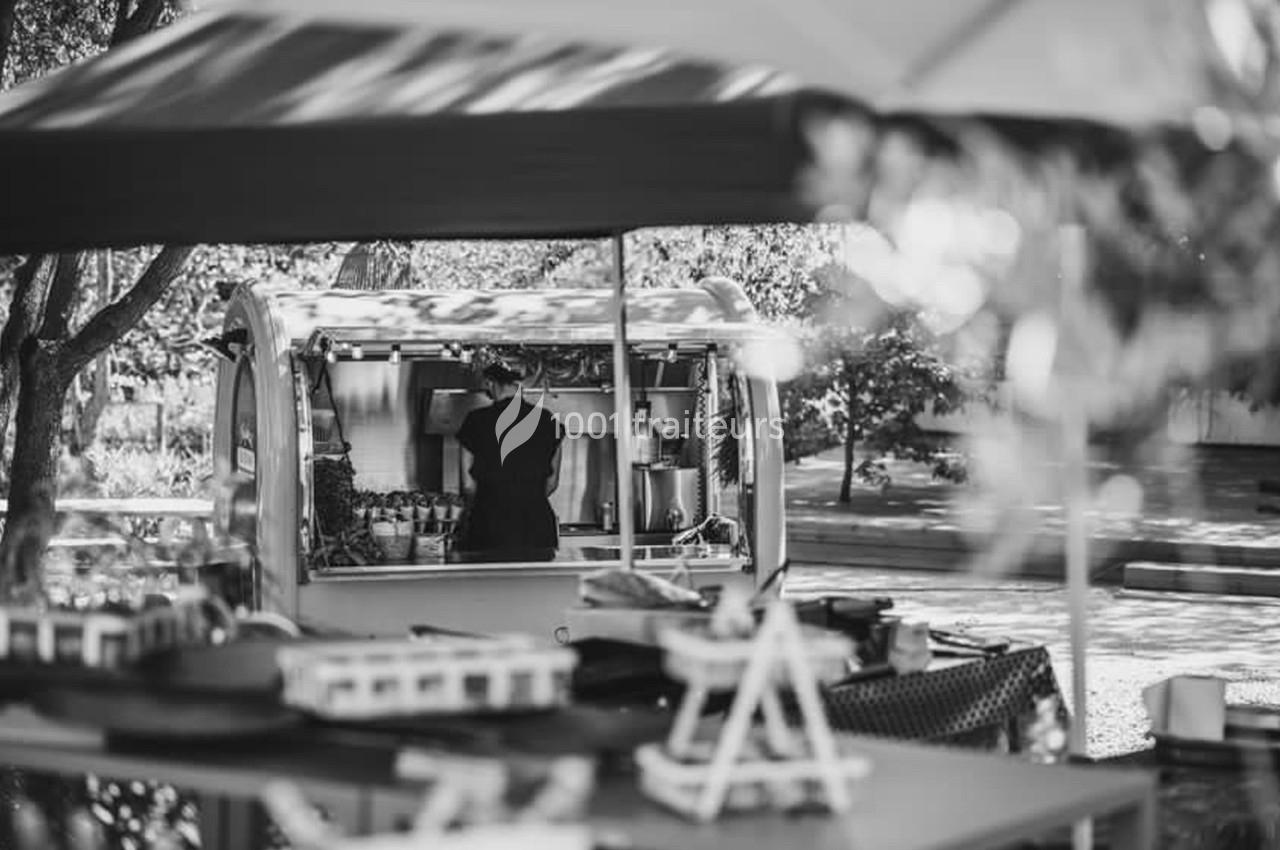 Un vendeur prépare des boissons dans une petite caravane aménagée, sous des parasols dans un espace extérieur ombragé.