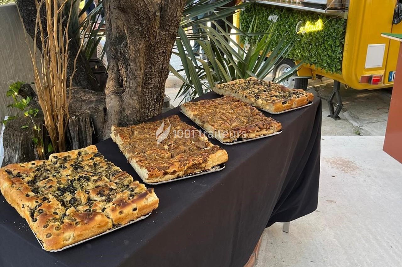 Quatre focaccias variées disposées sur une table noire en extérieur, près d'arbres et d'une remorque jaune.