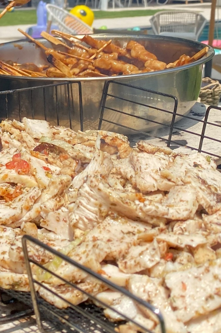 Viande grillée et brochettes en cuisson sur un barbecue en plein air, avec des tables en arrière-plan.