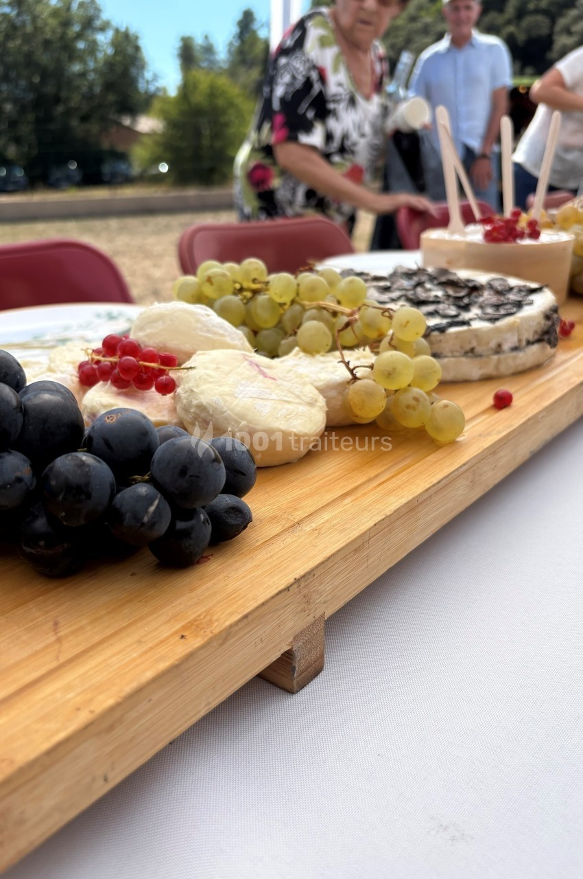 Plateau en bois avec fromages variés, raisins noirs et blancs, groseilles rouges, présenté sur une table en extérieur.