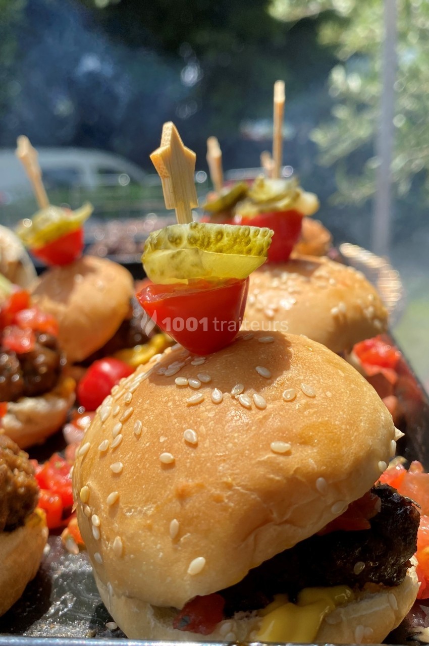 Mini burgers garnis de cornichons, tomates cerises et brochettes décoratives, présentés en extérieur ensoleillé.