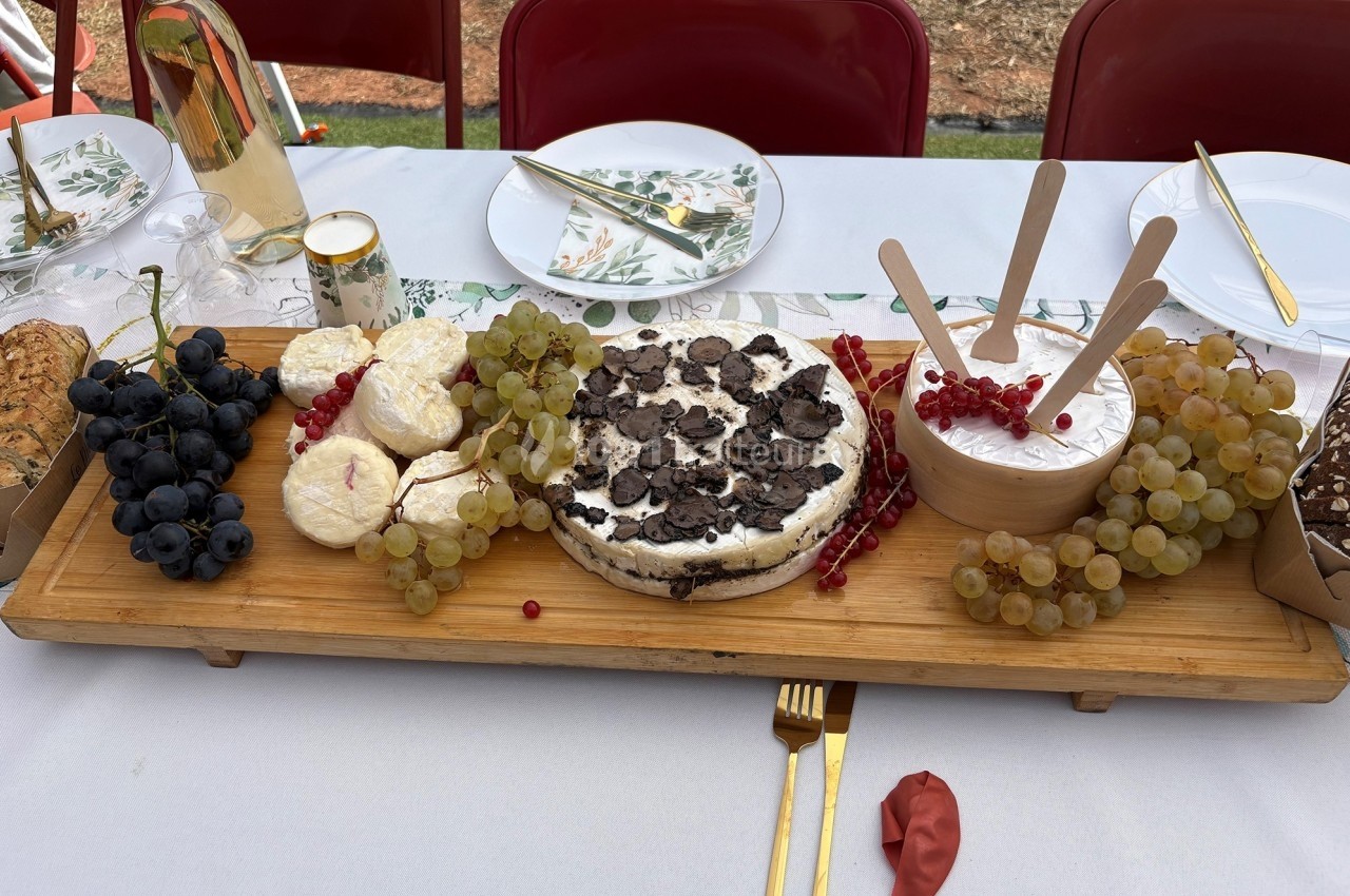 Plateau en bois avec fromages, raisins, truffes et groseilles, disposé sur une table dressée en extérieur.