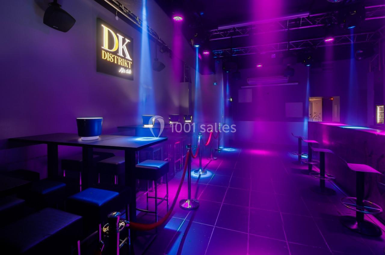 Salle de club avec éclairage violet et bleu, tables hautes, tabourets, cordons de séparation et logo mural lumineux.