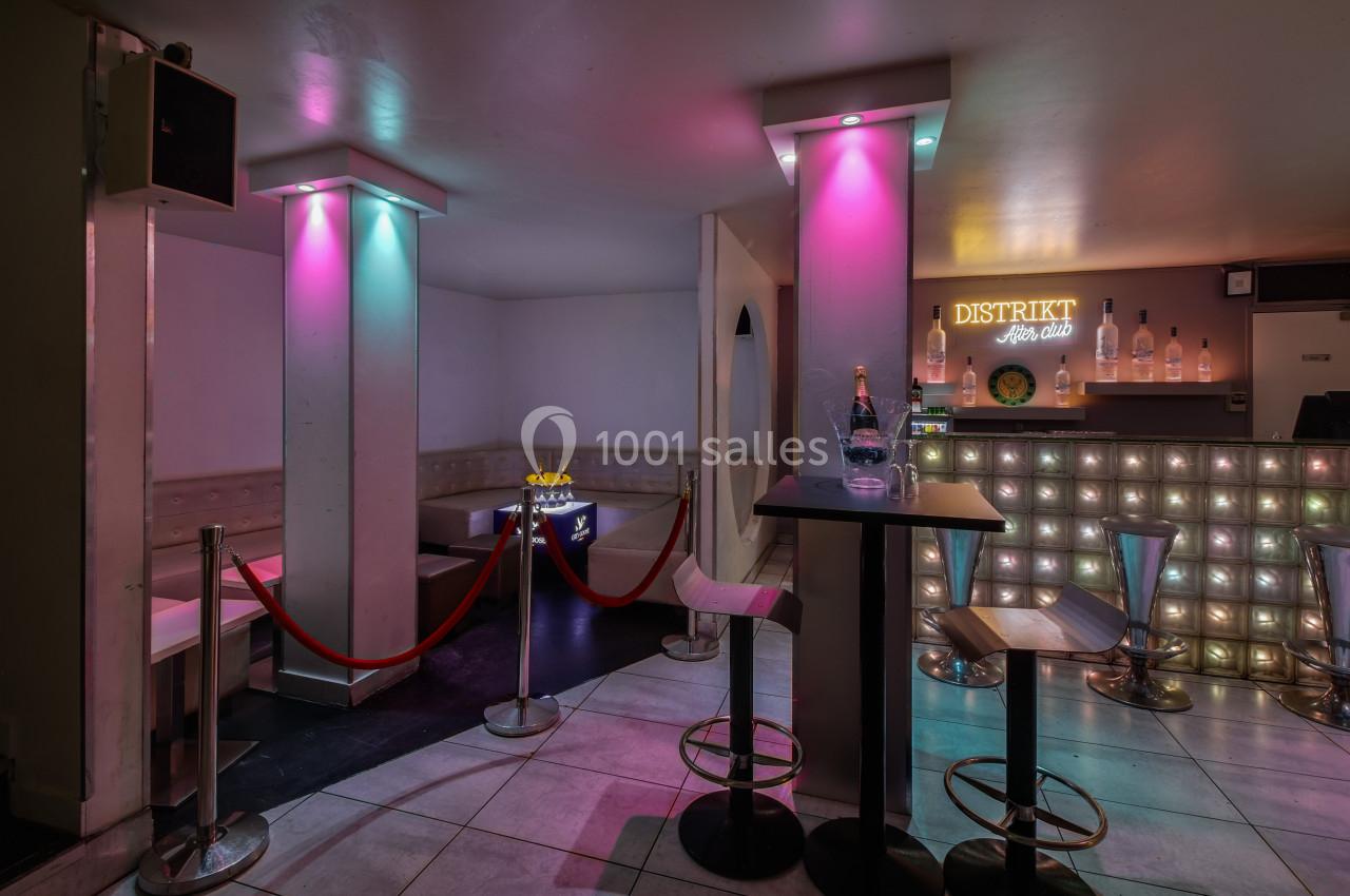 Intérieur d'un bar avec comptoir illuminé, tabourets, table haute et espace VIP délimité par des cordons rouges.