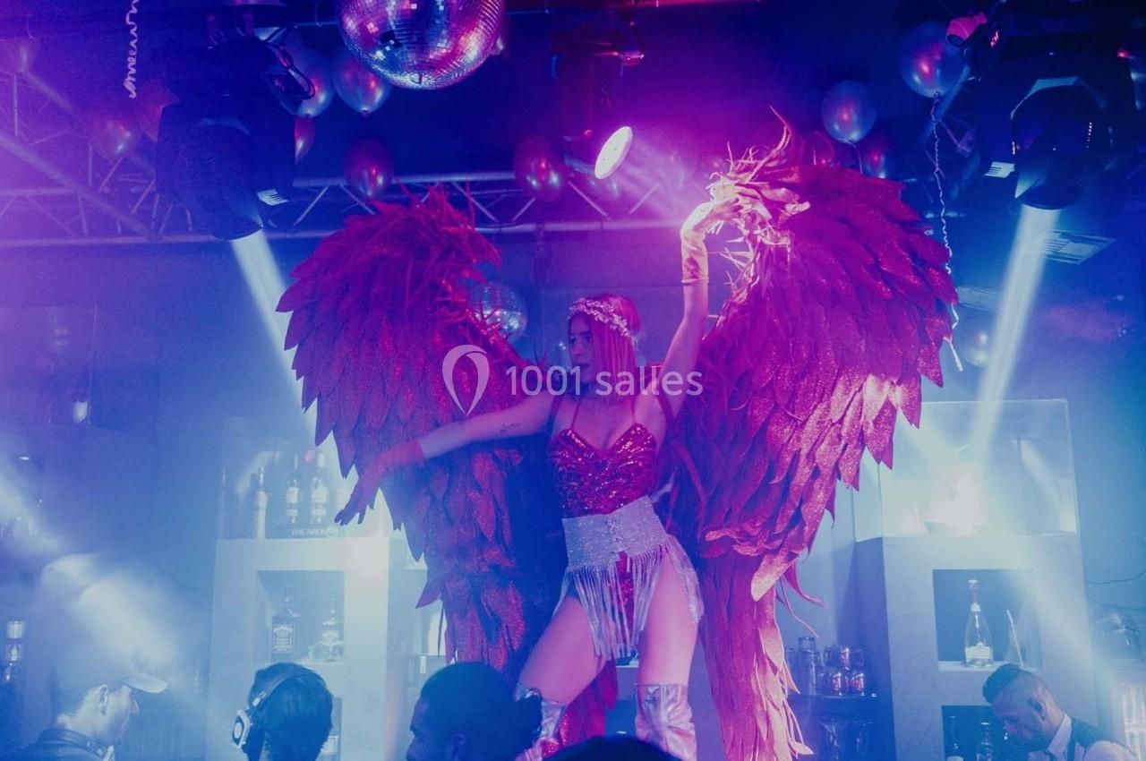 Une danseuse en costume rouge avec de grandes ailes, éclairée par des lumières colorées dans une ambiance de club.