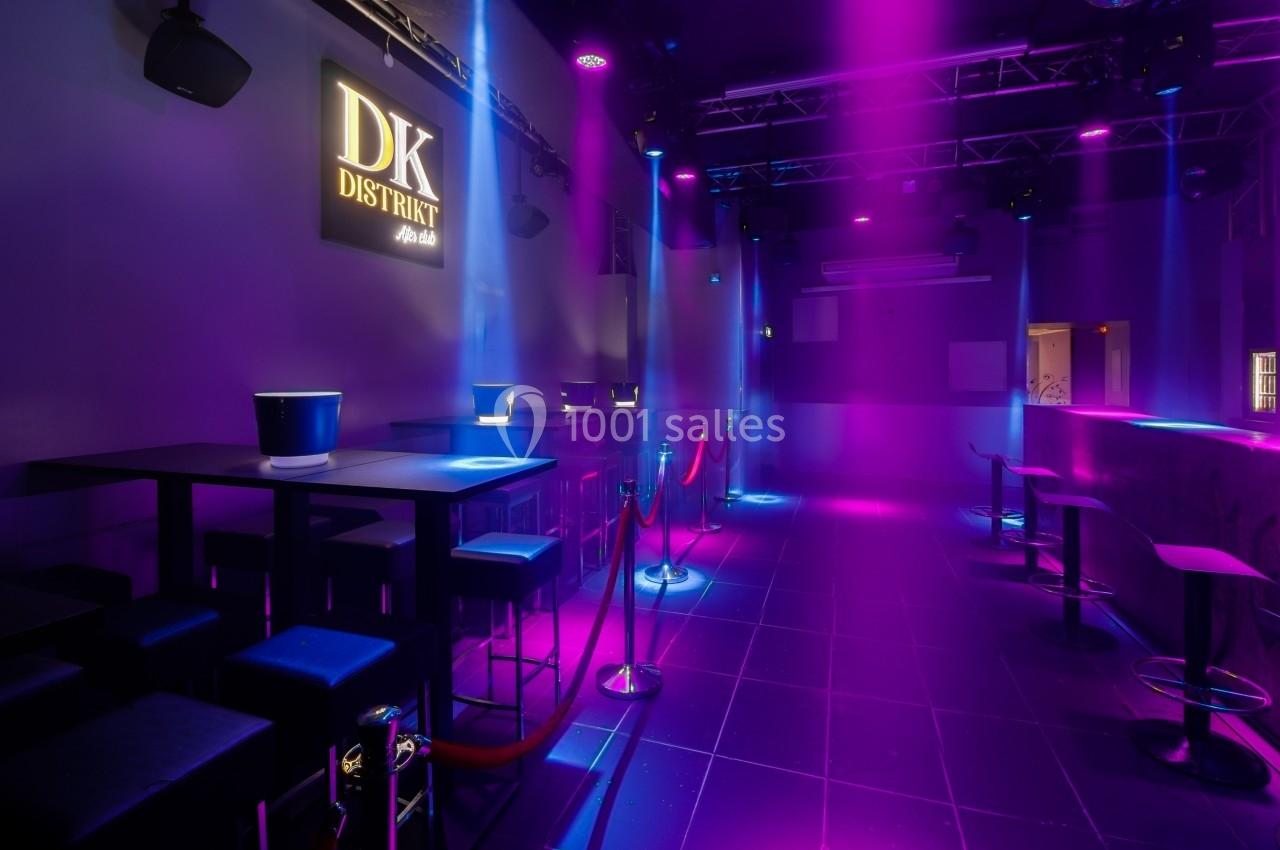 Salle de bar moderne avec éclairage violet et bleu, tables hautes, tabourets et cordons de séparation rouges.