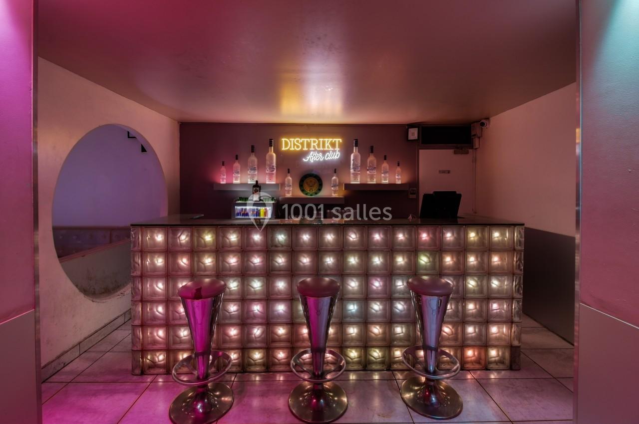 Bar avec comptoir en briques de verre illuminées, trois tabourets hauts et enseigne lumineuse ’Distrikt’ en arrière-plan.