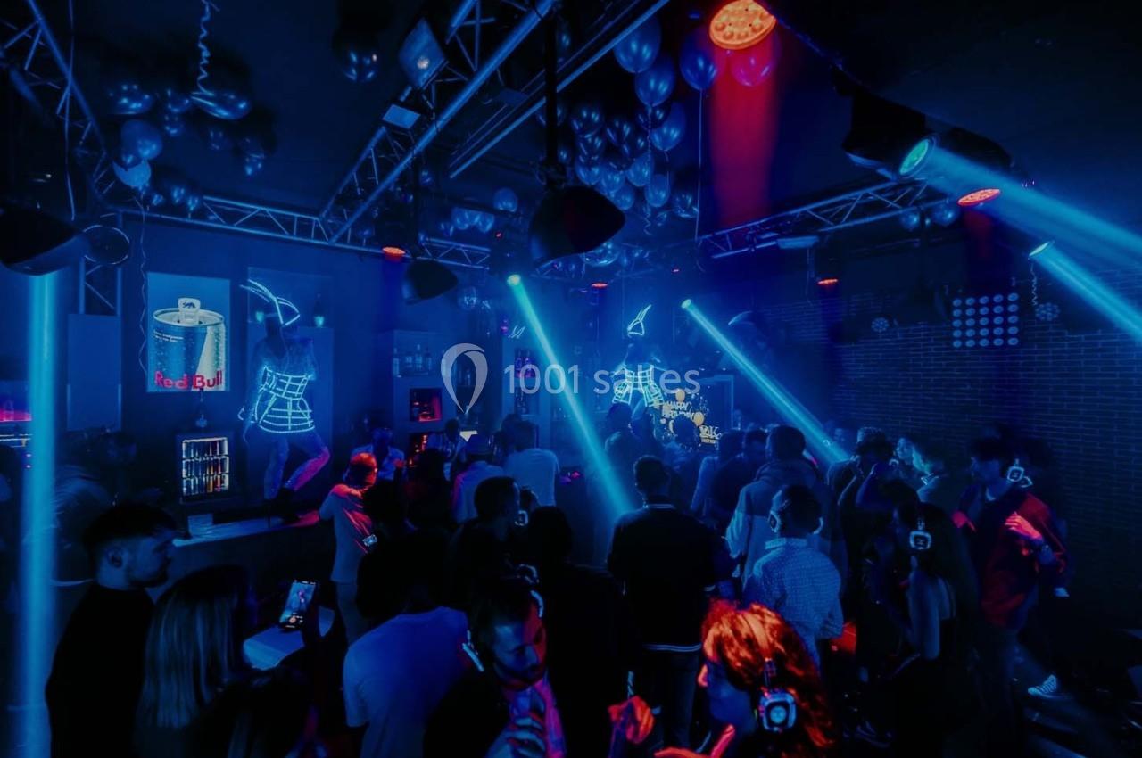 Foule dans une salle de concert sombre avec éclairages bleus, DJ sur scène et ambiance festive.