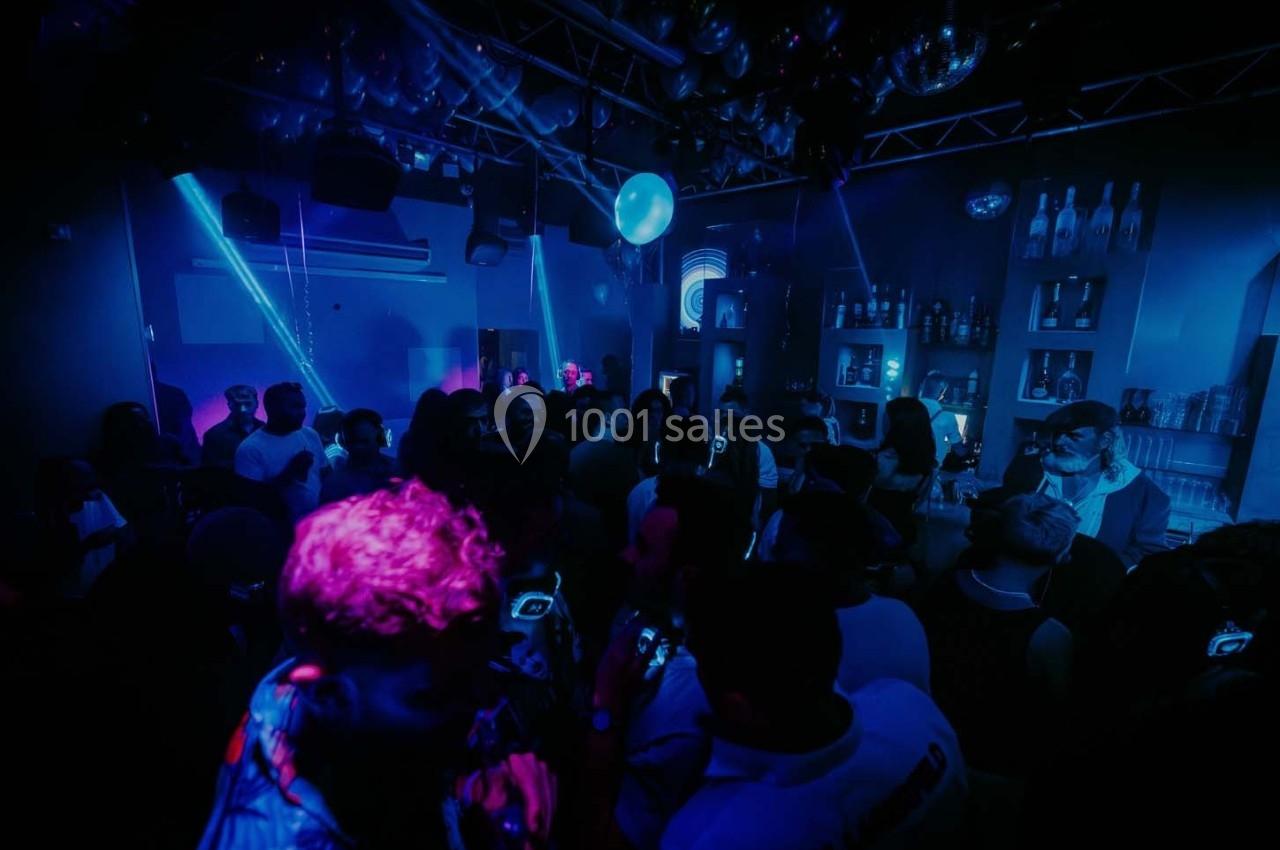 Foule dans une boîte de nuit sombre éclairée par des lumières bleues et violettes, avec des étagères de bouteilles en…