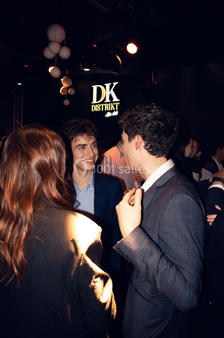 Trois personnes discutant dans un lieu sombre avec une enseigne lumineuse ’DK Distrikt’ en arrière-plan.