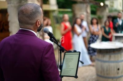 Un homme en costume violet parle au micro lors d'un événement en plein air, avec des invités flous en arrière-plan.