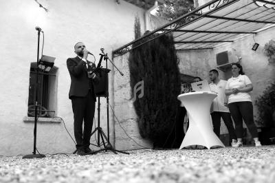 Un homme en costume parle au micro sur une petite scène extérieure, devant un public et près d'une table haute.