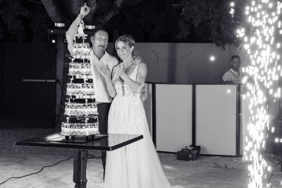 Un couple souriant coupe une pièce montée lors d'une célébration en extérieur, avec des étincelles en arrière-plan.