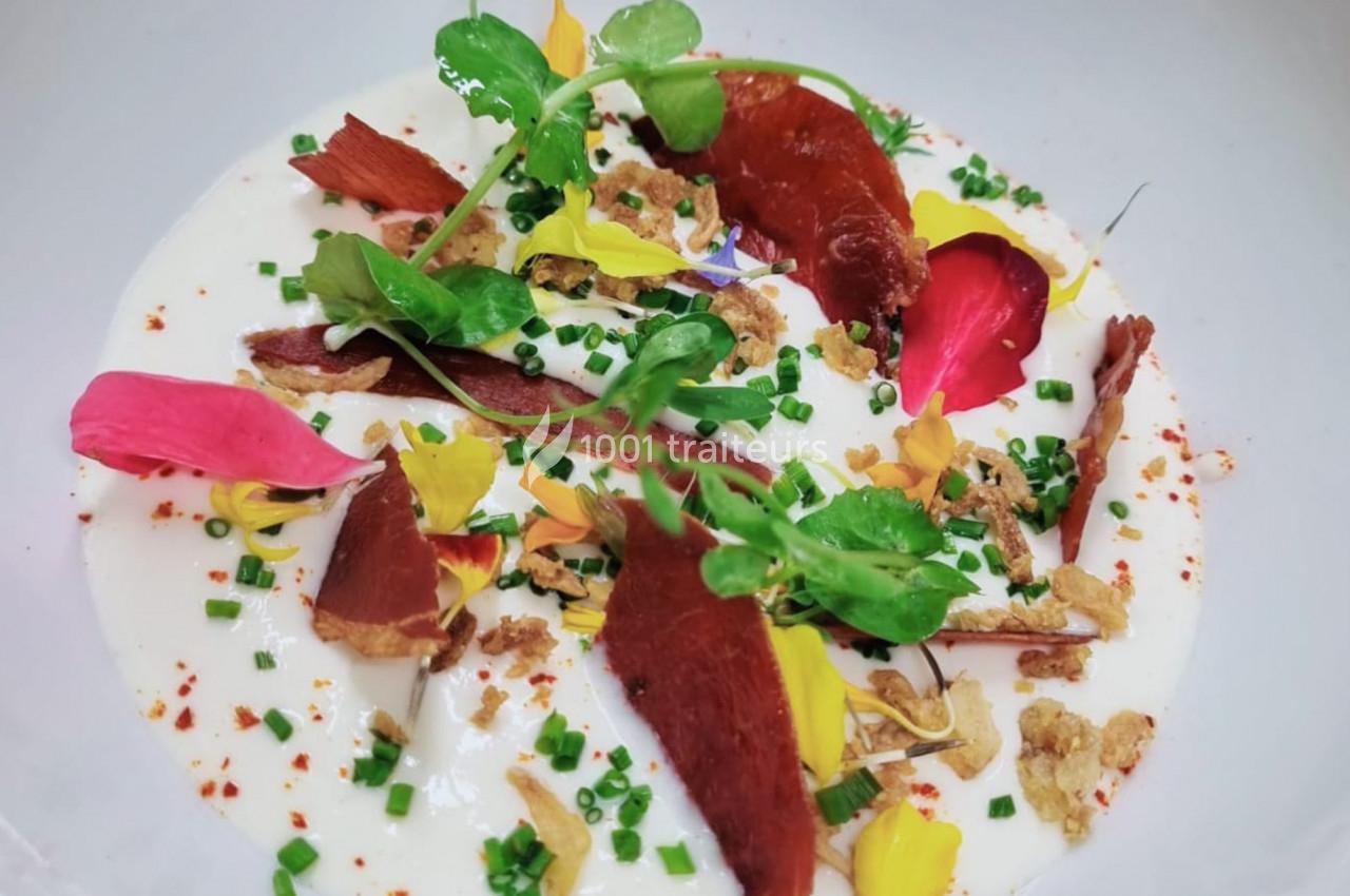 Assiette de velouté blanc garnie de fines tranches de viande séchée, fleurs comestibles, herbes et croûtons.