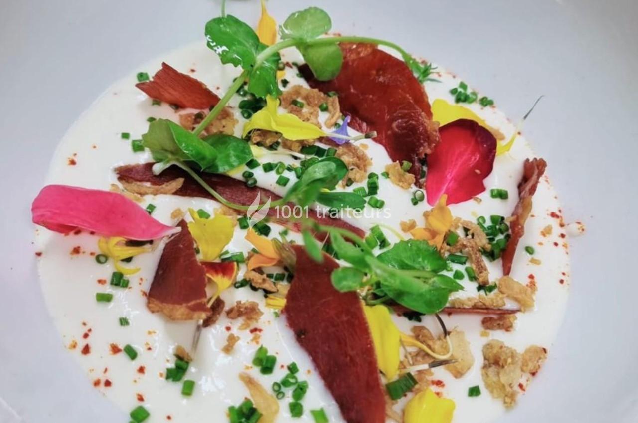 Assiette garnie de crème blanche, pétales de fleurs colorées, herbes fraîches, morceaux de viande séchée et croustillants.