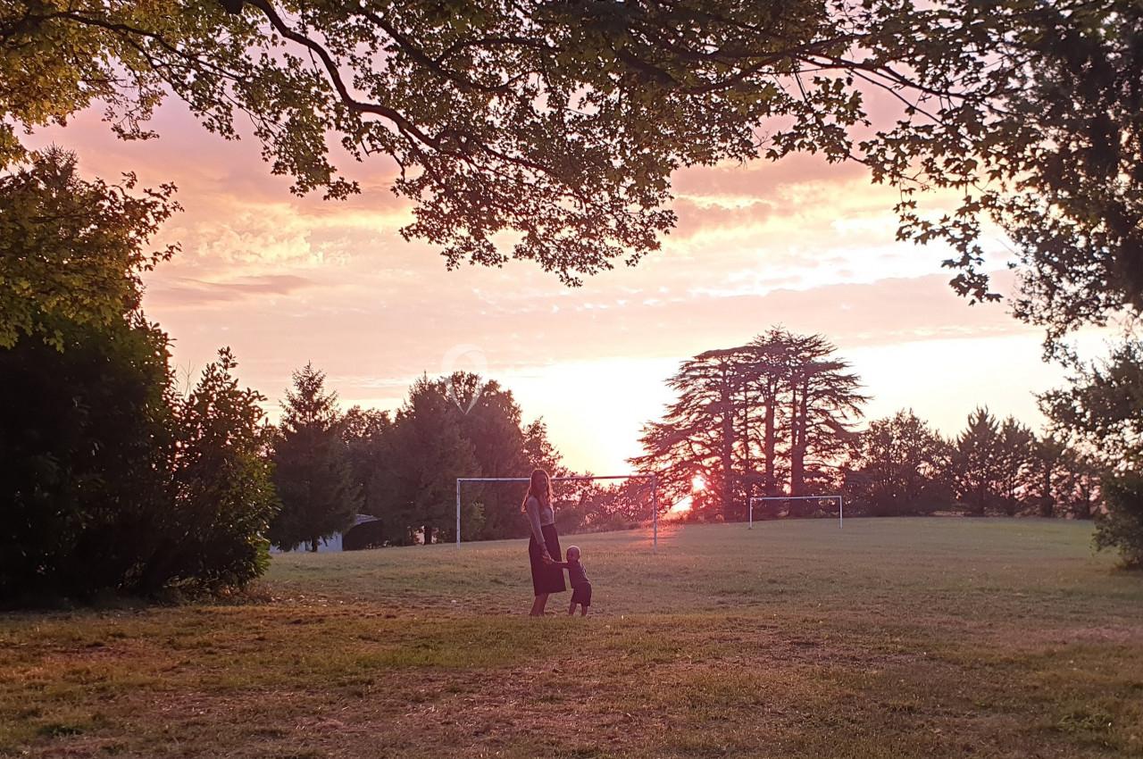 Une femme marche avec un enfant dans un parc au coucher du soleil, entourés d'arbres et de pelouses.
