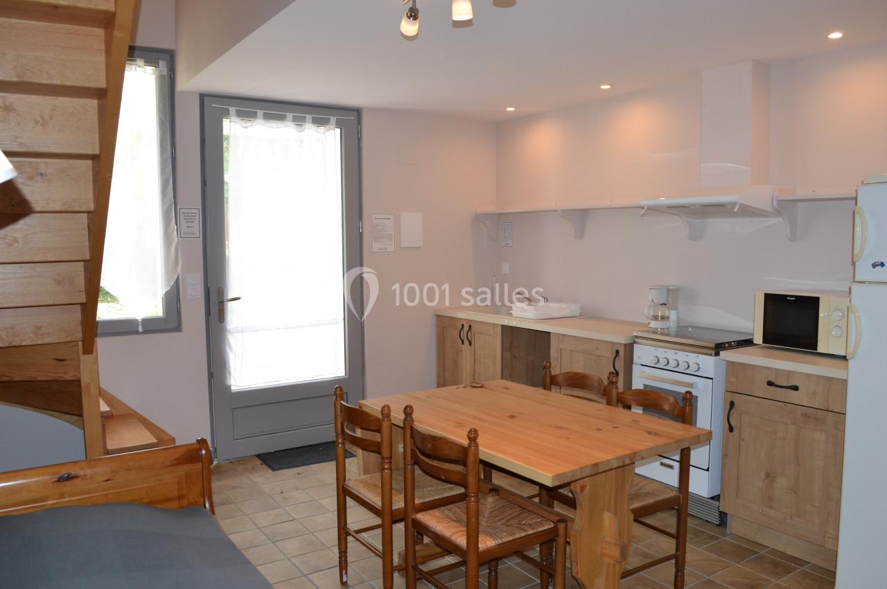 Cuisine ouverte avec table en bois, chaises, réfrigérateur, cuisinière, micro-ondes et escalier en bois visible à gauche.