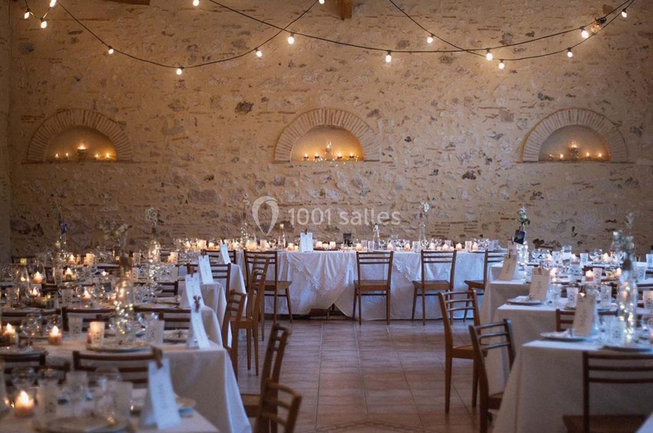 Salle de réception en pierre avec tables dressées, chaises en bois et guirlandes lumineuses suspendues.