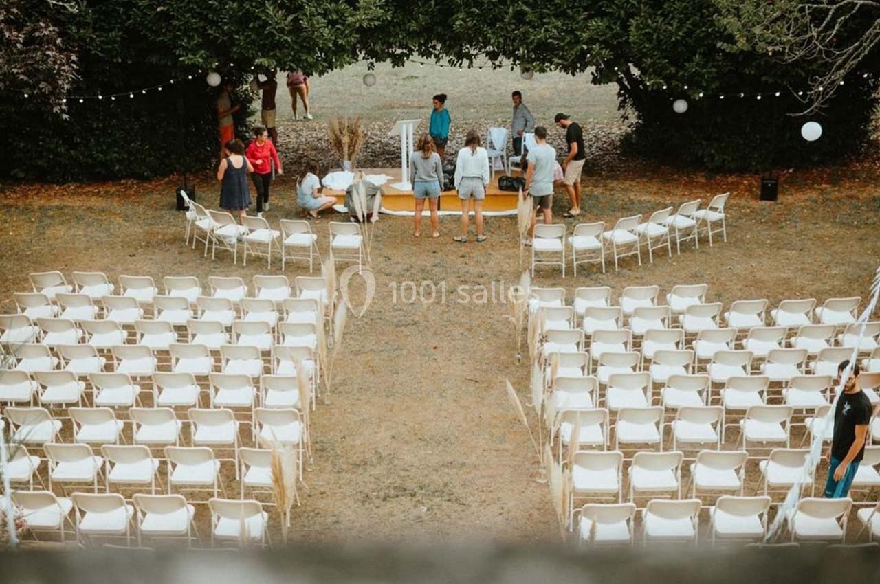 Chaises blanches disposées en rangées en extérieur, avec quelques personnes préparant une scène sous des arbres.