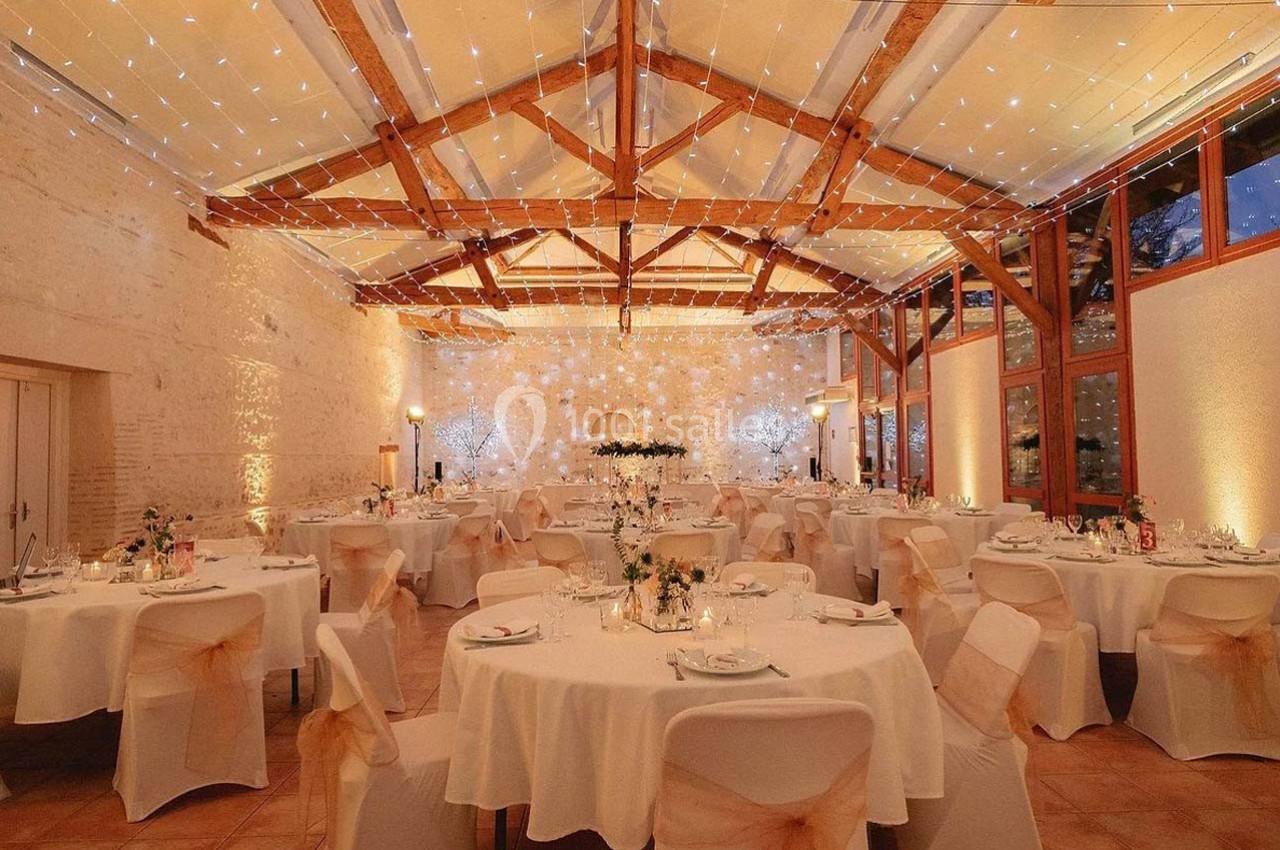 Salle de réception décorée avec des tables rondes, nappes blanches, chaises ornées et guirlandes lumineuses suspendues.
