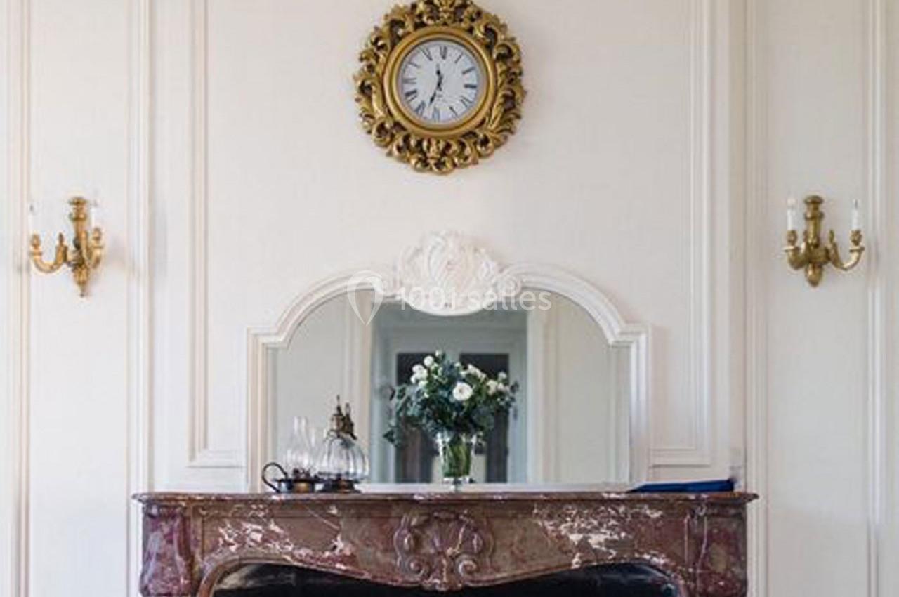 Horloge murale dorée au-dessus d'une cheminée en marbre rouge avec un miroir et un vase de fleurs.