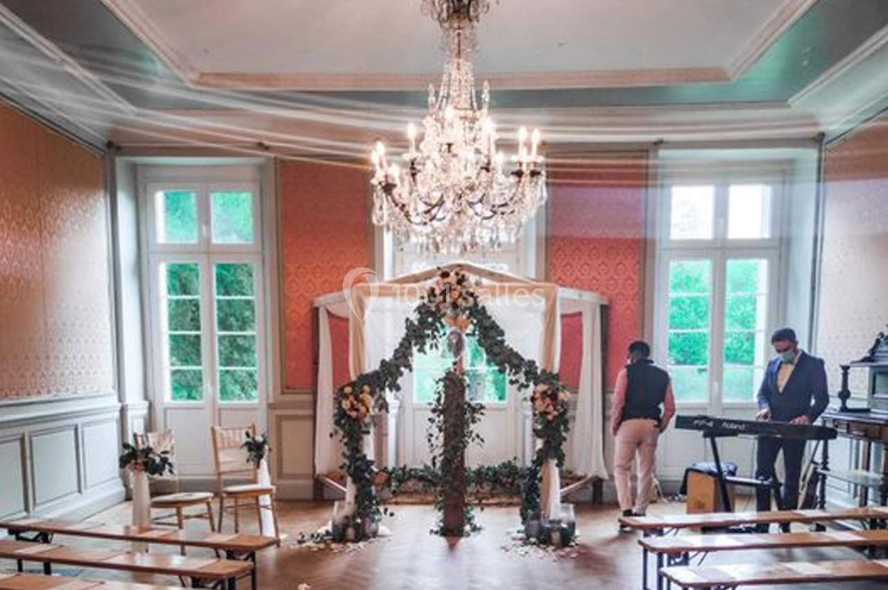 Salle décorée pour une cérémonie avec arche fleurie, chaises alignées et deux personnes préparant l'ambiance musicale.