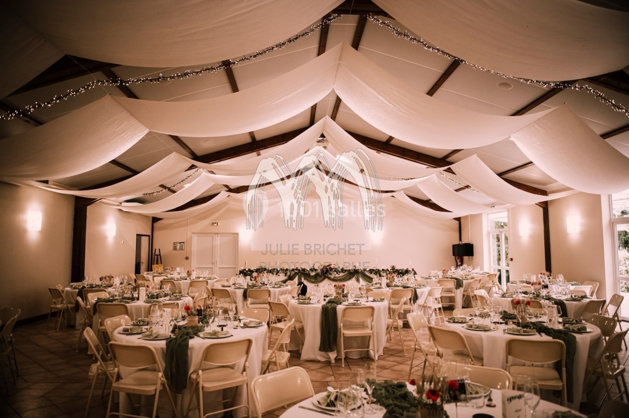 Salle de réception décorée avec des tables rondes dressées, nappes blanches et guirlandes lumineuses suspendues.