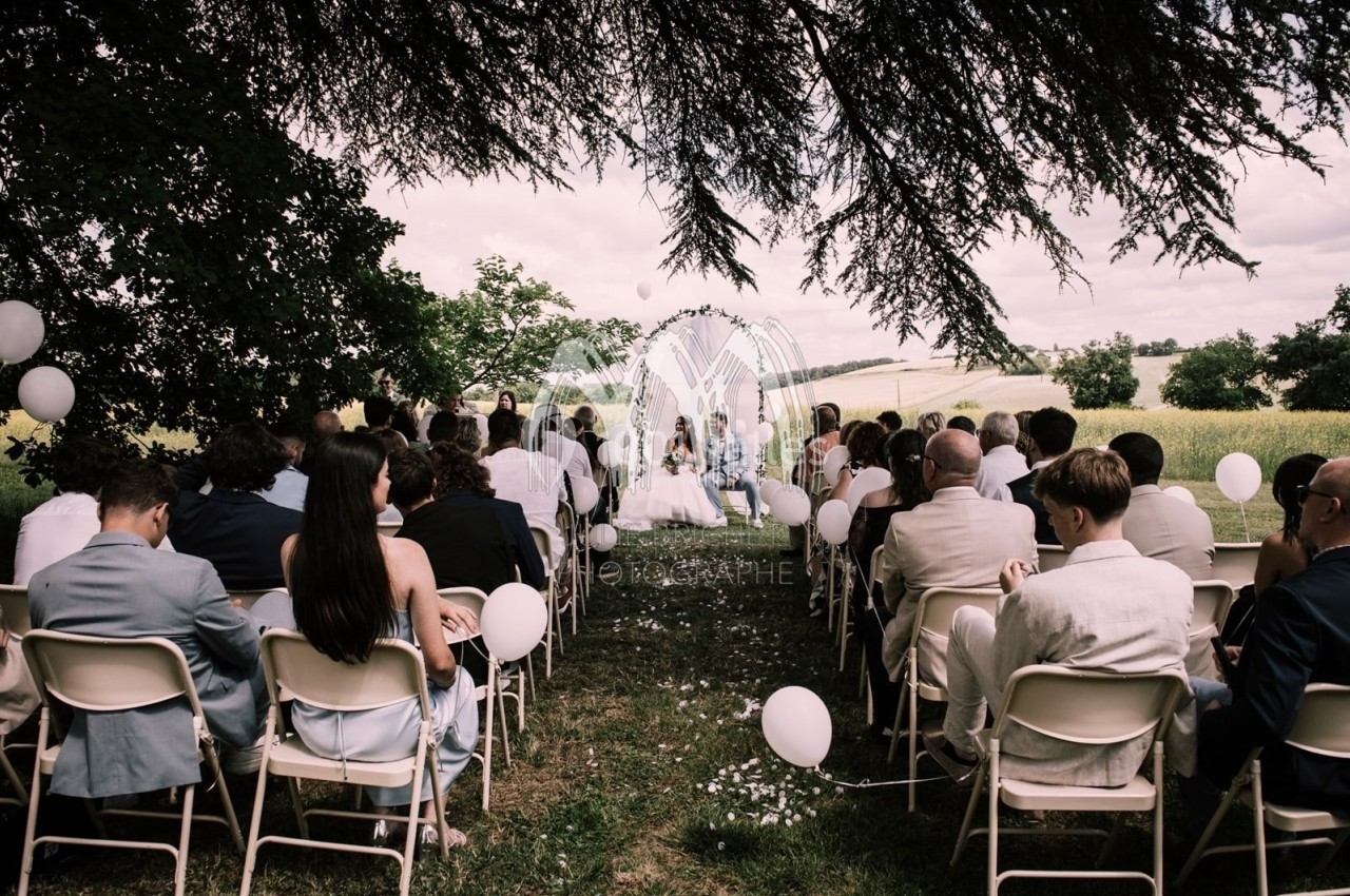 Cérémonie en plein air avec des invités assis sous des arbres, face à un couple devant un paysage champêtre.