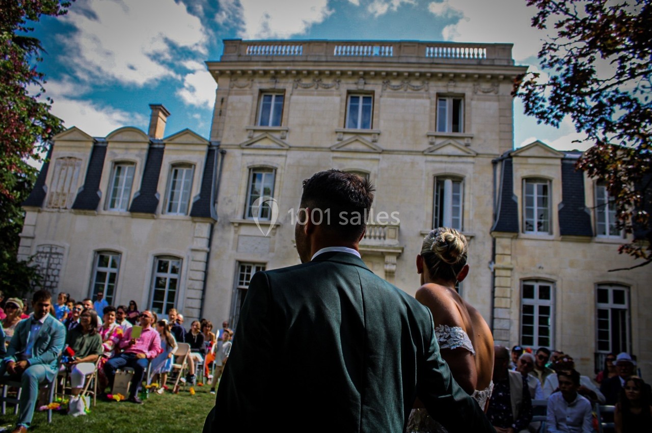 Un couple de mariés de dos face à des invités assis dans le jardin d'un grand bâtiment en pierre.