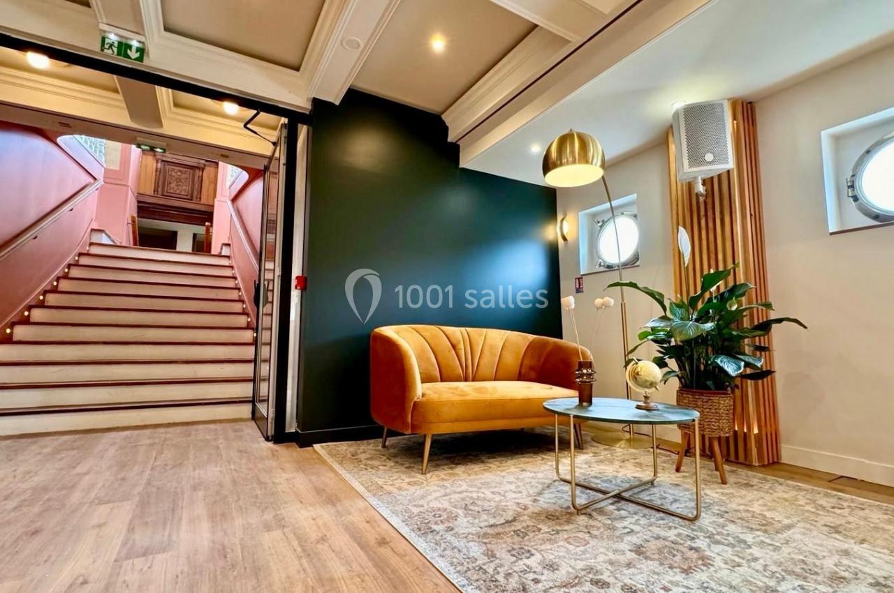 Salon lumineux avec canapé jaune, table basse en verre, plante verte et escalier en arrière-plan.