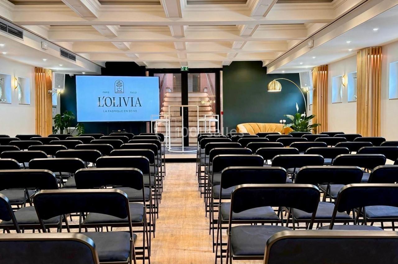 Salle de conférence lumineuse avec rangées de chaises noires, écran de présentation et décor moderne.